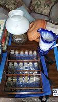 Wooden spice rack filled with 18 small glass bottles with stoppers, Burleigh vase, terra cotta baker, blue pitcher, and pottery oil lamp on a wooden surface