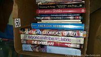 Books stacked in a box, titles visible including 'The Dresden Party Needlework', '5000 Years of Textiles', 'Fiberarts Design Book Four', 'American Needlework', 'America's Glorious Quilts', 'Beyond Craft: The Art Fabric', 'The Dyer's Art'. Condition shows use wear.