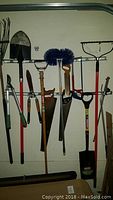 Wide view showing various hanging yard tools including shovels, saws, broom, pruners, and rakes hung on a wall rack.