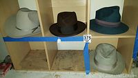 Four vintage men's hats placed in wooden cubby shelf: a light gray Western style, dark brown classic felt, teal wool, and light gray felt hat