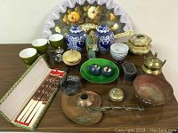 Full view of the lot on table showing chopsticks in box, porcelain cups, ginger jars, cloisonne items, fan, pottery figure, small containers, and solid copper candle holder.