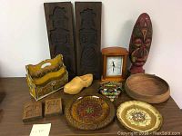 Full lot view showing carved wooden plaques, painted mask, bowls, decorative plates, magazine holder, carved animal blocks, wooden clog, and chalet clock arranged on table.