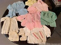 Lot of vintage baby sweater sets arranged on a table including blue, pink, green, cream, and yellow sweaters with matching bonnets and booties.