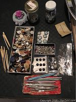 Top view of various buttons in a large box, smaller boxes of buttons, pearl-headed pins in a box, and several metal crochet hooks laid out in a red pouch.