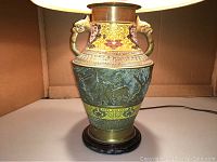 Front view of the lamp illuminated showing detailed cloisonne enamel artwork with floral designs and raised animal figures on brass base with dual handles.