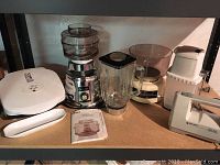 Full view of all items in the lot on shelf including Osterizer Deluxe blender with food processor accessory, George Foreman grill, Swing Away ice crusher, food scale, and Black & Decker hand mixer without beaters.