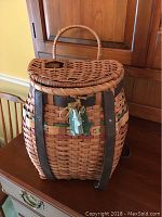 Front view of the woven fishing basket showing hinged lid, leather straps, and carrying handle with attached decorative tag.