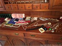 Various costume jewelry pieces arranged on wooden dresser surface showing necklaces, bracelets, pins, and earrings including boxed items and multi-strand pearl necklaces.