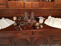 Wide view of the lot items on wooden cabinet including baby dresses, dressing table items, glass tumblers, and an apron.