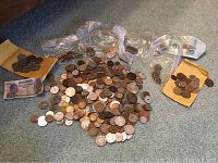 Top-down view of a large quantity of assorted foreign coins, some in small plastic bags and some loose on a carpeted floor.
