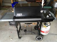 Front view of Weber gas grill with propane tank, showing the grill body, wheels, side shelf, and lower storage shelf with grilling tools.