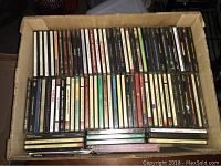 Top-down photo of open cardboard box filled with stacked music CD jewel cases in various colors and text on spines.