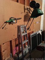 Green garden spreader, aluminum step ladder, and green electric hedge trimmer hung on pegboard.