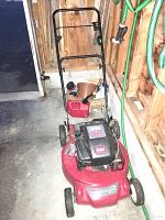Front and side view of red Toro gas lawn mower with black handle and wheels, sitting on concrete floor with a green garden hose in the background.