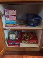 Photo inside wooden cabinet shelves showing various boxed games including Pictionary and Buzz Word, some items in blue crate.