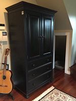 Full frontal view of the tall black solid pine armoire with two closed doors, three drawers, standing on hardwood floor next to a guitar.