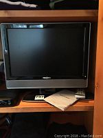 Front view of the 19 inch Insignia LCD TV on shelf with remote and manual placed in front.