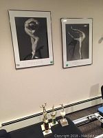 Two framed black and white ballet prints on the wall with four volleyball trophies on the heater base below them.