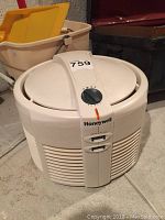 Whole view of white Honeywell air purifier with dial control, mop bucket and mop handle behind it.
