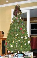 Photo showing the full artificial Christmas tree decorated, standing indoors with an angel tree topper and various ornaments, though decorations are not included in the sale.