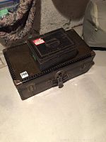 Overall view of a large black metal trunk with rivet detailing and a smaller matching metal box placed on top, showing the full lot.