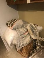 Photo shows a pile of pillows in clear plastic bags sitting on a woven laundry hamper next to a white floor fan, also visible are striped and polka dot curtains in background.