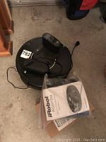 Photo showing black iRobot Roomba vacuum robot on floor with attached charging dock and power cord, owner's manual in plastic bag visible.
