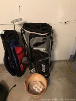 Photo shows two golf bags; one Cooper brand in black, silver, and white, the other red and blue. A wooden bucket filled with golf balls is placed in front. A putter is in the red and blue golf bag.