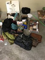 Photo shows assortment of women's items including purses, scarves, jewelry boxes, skincare products, and a white electric device arranged on a flat surface.