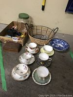 Overview shot showing six assorted porcelain tea cups with matching saucers in floral patterns and gold trims, blue and white bowl, metal basket, and glass jar with green lid on concrete floor.