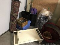 View of the lot showing the rolled area rug, white metal bed tray, black umbrella stand with umbrellas, blue container, and multiple wicker baskets of various sizes and styles.