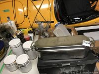 Photo of a stainless steel salmon roaster with lid, multiple white-lidded glass jars, measuring jug, and other kitchen utensils surrounding a yellow car door in background.