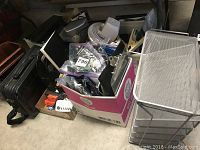 Overview of lot showing black leather briefcase on left, pink and white box of assorted office supplies including cords and hardware, white metal mesh shelving unit on right, small white fan, and parts on floor including casters and tools.