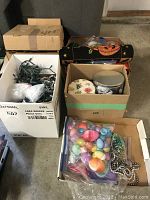 Three cardboard boxes containing assorted Christmas ornaments, globe string lights, and other decoration materials in plastic bags.