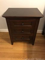 Mahogany cherry-finished nightstand against white wall on wood floor, showing three drawers and metal knobs.