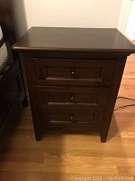Straight-on view of the actual nightstand showing three drawers, round metal knobs, mahogany construction and cherry finish on hardwood floor