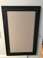 Image of a planner or bulletin board with a beige cloth surface surrounded by a black wooden frame, including metal side hinges and a handle.