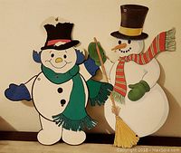 Two wooden snowman figures with hats and scarves on a flat surface against a beige wall.