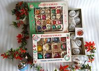 Overhead photo showing two boxes of vintage glass Christmas tree ornaments, white crochet ball ornaments, silver reflective ornament, clear glass ornament with red design, and multiple artificial red poinsettia floral picks.