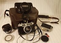 Camera with leather carrying case, flash unit, accessory strap, lens ring, cleaning brush, and lens cap arranged on cloth background.