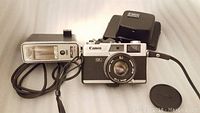 Photo shows Canon Canonet QL camera with attached lens, a Canon flash with cable, and a black carrying case positioned behind the camera.