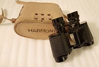 Photo showing the black Harmony 8x30 binoculars along with the tan carrying case labeled 'HARMONY'.