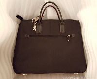 Front view of the black lady's handbag/briefcase showing brown handles, a front zippered pocket, and a silver-tone charm attached to one handle.
