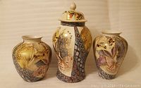 Photo showing a set of three hand-painted decorative vases from the late 1980s. Two vases are rounded and bulbous; the third is a taller cylindrical vase with a lid featuring a knob. All have consistent color schemes and design motifs of butterflies and flowers.
