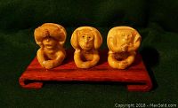 Photo of three wise monkeys figurines carved from light-colored material, mounted on a reddish wooden base showing 'see no evil, hear no evil, speak no evil' poses.