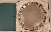 Top-down view of round Birks silver plated tray inside its blue box with tissue lining. Tray features scalloped edge and central etched scroll floral pattern.