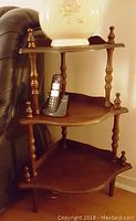 3-tier wooden corner shelf with dark finish shown holding a vase and cordless phone, designed with scalloped edges and turned spindle supports.