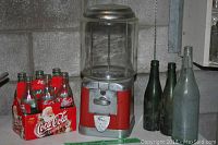 Red and silver Beaver gum ball machine next to Coca-Cola six-pack glass bottles and three large old glass bottles on table