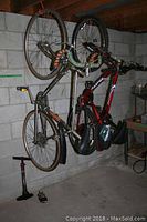 Photo showing two bicycles hanging vertically by their front wheels on wall hooks in a basement, one silver Apollo speed bike and one red Huffy mountain bike. Floor pumps are on the floor below.