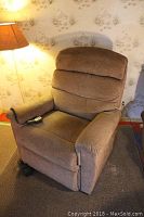 Brown La-Z-Boy electric recliner chair shown from the front-left side with remote control on armrest and lamp beside it.
